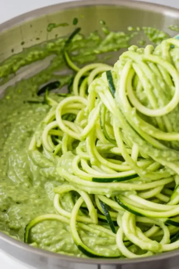 Healthy zucchini noodles being mixed in a creamy avocado sauce inside a stainless steel pan